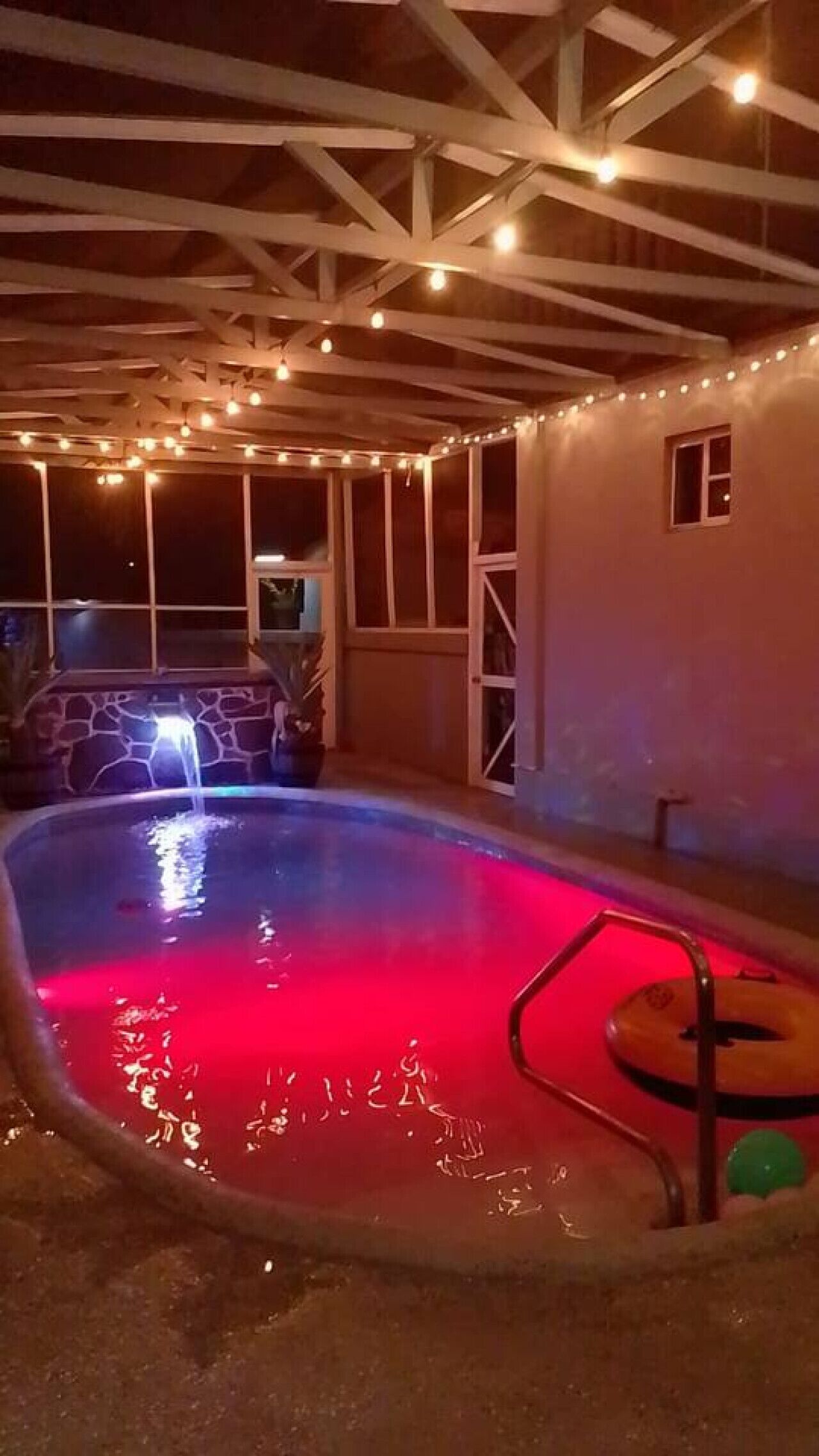 An outdoor swimming pool with a red light, surrounded by a covered area with string lights.