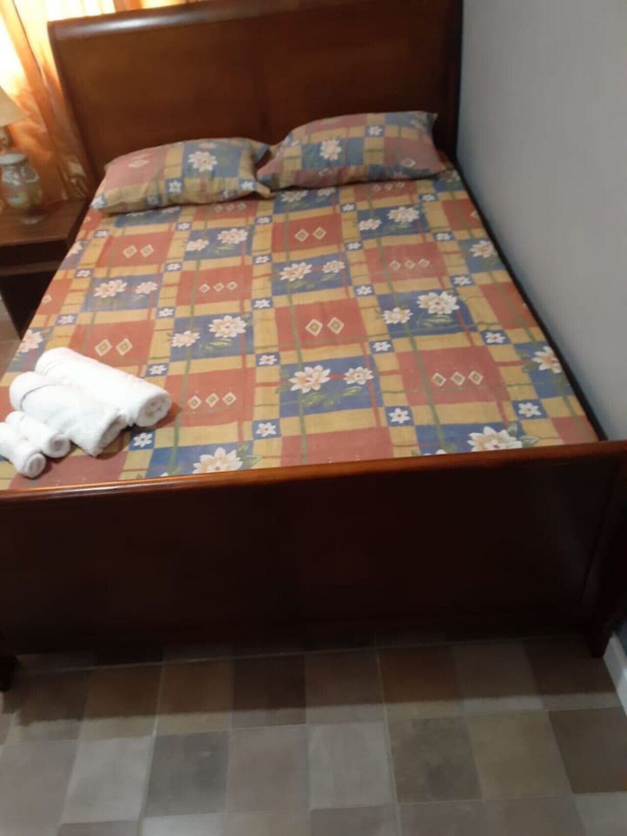 Un letto con copriletto a motivi floreali, due cuscini e una coppia di asciugamani arrotolati su un lato.
