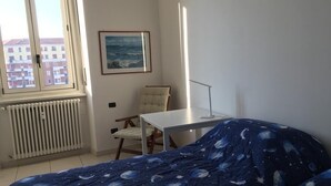 Quarto com cama, cadeira, escrivaninha, radiador, janela e um quadro na parede.