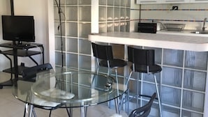 Uma cozinha moderna com armários brancos, painel de vidro atrás do fogão e um pequeno espaço de jantar com mesa de vidro e cadeiras pretas.
