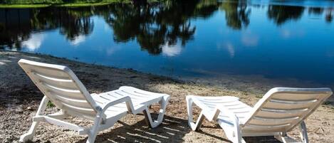 Deux fauteuils blancs sont placés face à un lac paisible, avec un ciel dégagé et une végétation luxuriante en arrière-plan.