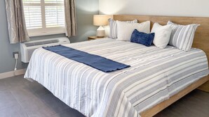 A neatly made bed with striped bedding and a blue throw blanket, a bedside lamp, and a window with curtains. - ADA Poolside King Studio (Marco Island)