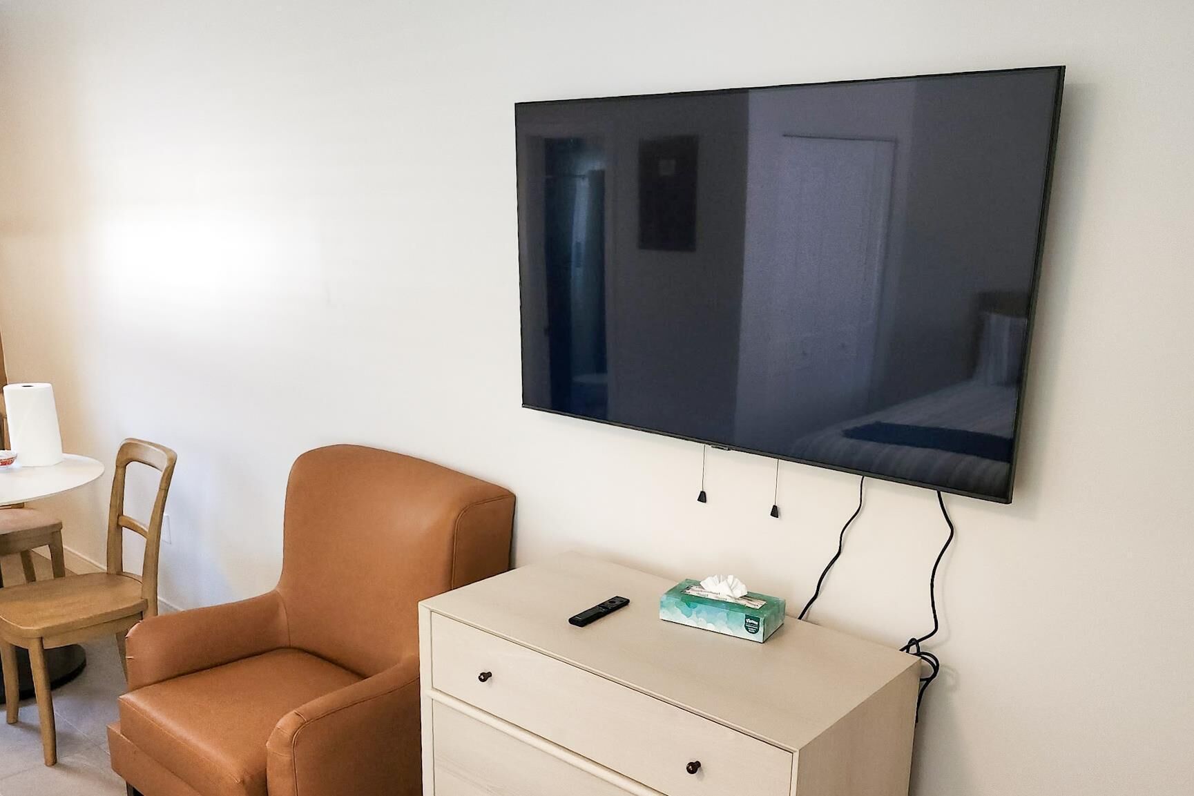 A room with a large flat-screen TV mounted on the wall, a brown leather chair, a small white desk with drawers, and a remote control on the desk.