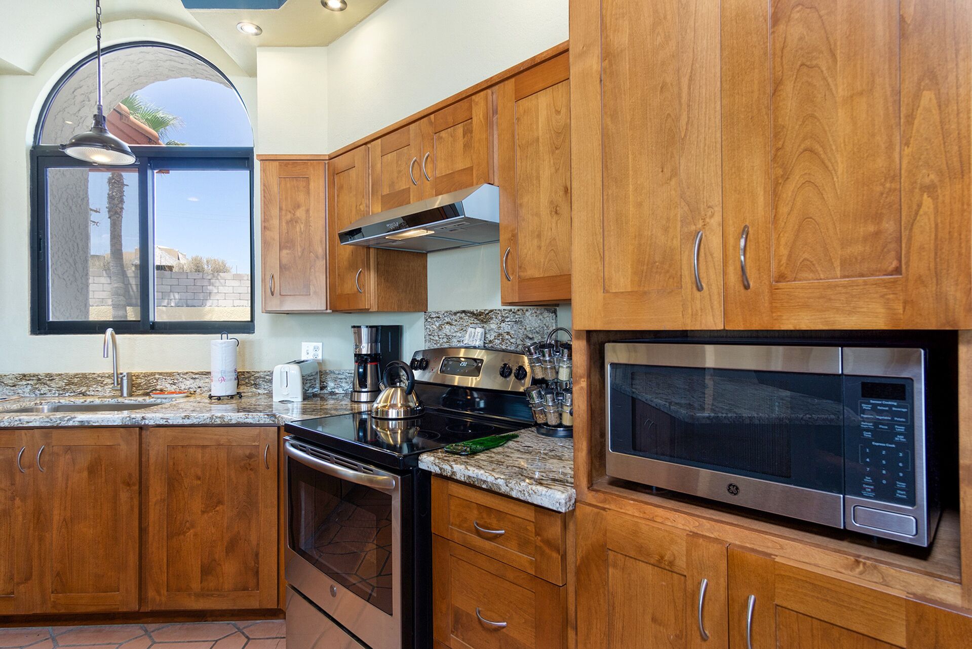A kitchen with wooden cabinets, a stainless steel stove, a microwave, and a window with a view of a palm tree and buildings.