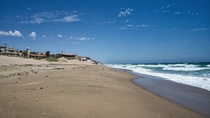 Une plage avec un rivage sablonneux, quelques bâtiments en arrière-plan et les vagues de l’océan qui se rapprochent du rivage.