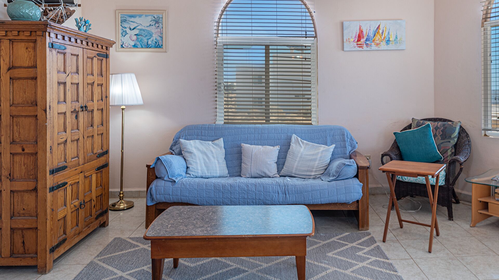 A living room with a blue sofa, a wooden cabinet, a floor lamp, and a window with blinds.