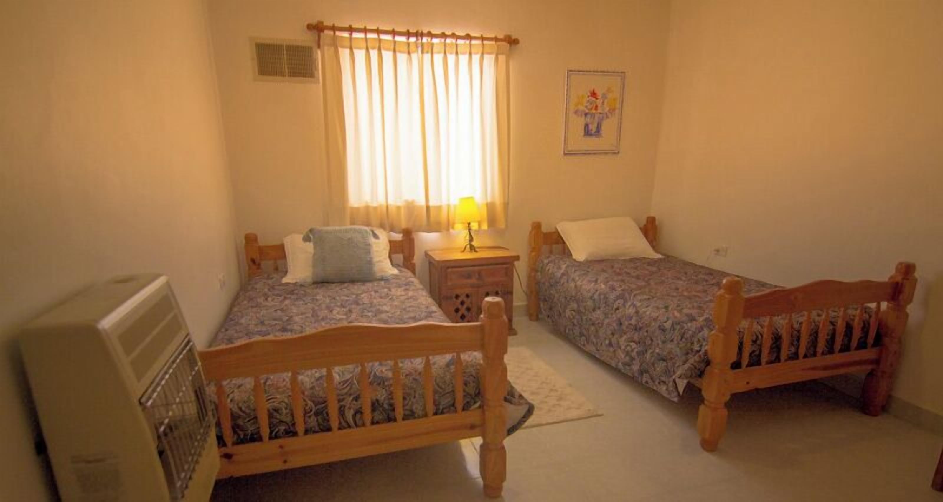 A bedroom with two wooden beds, a bedside table with a lamp, a radiator, and a window with curtains.