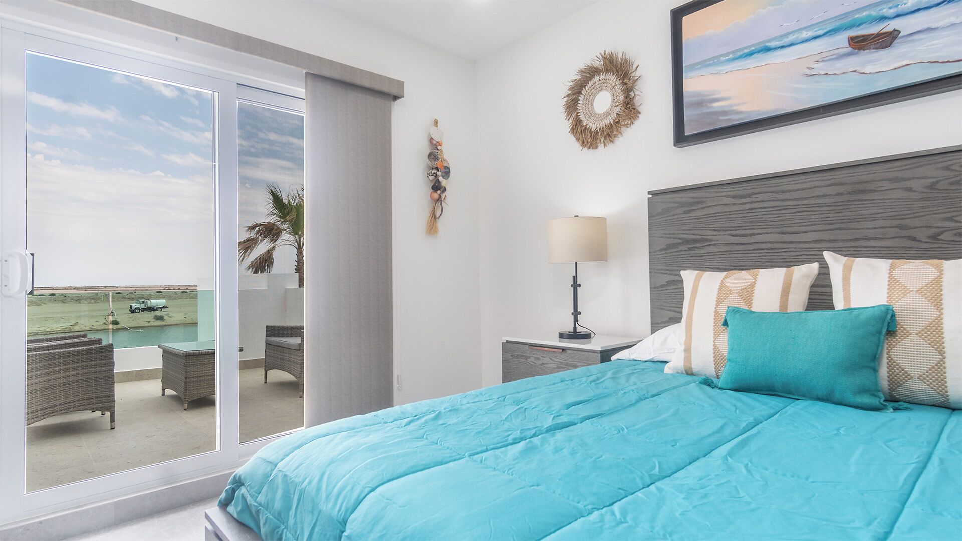 Quarto com cama grande, mesa de cabeceira, luminária e vista para a praia através de uma porta deslizante.