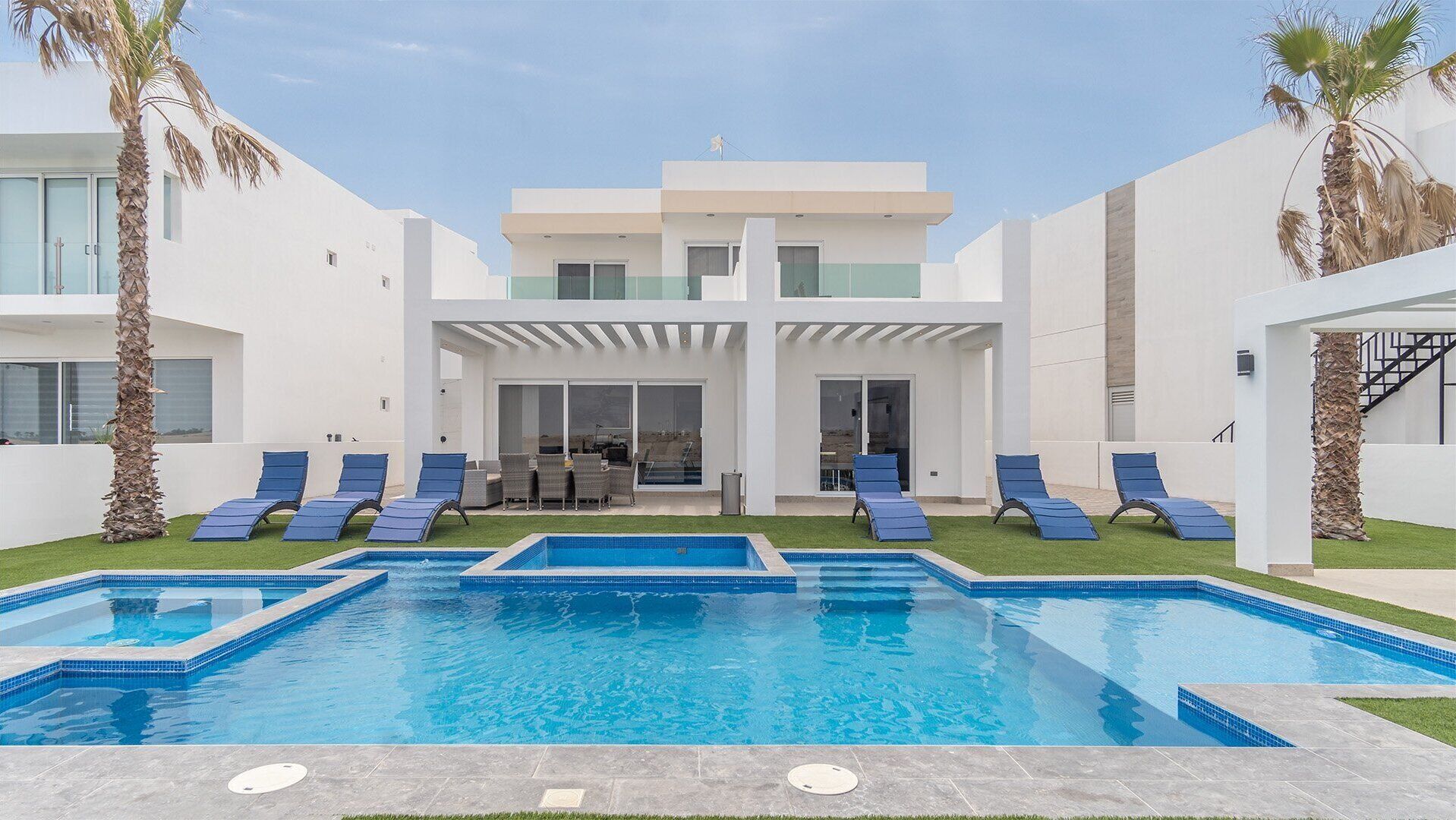 Ein modernes, zweistöckiges Haus mit Swimmingpool, Liegestühlen und Palmen.