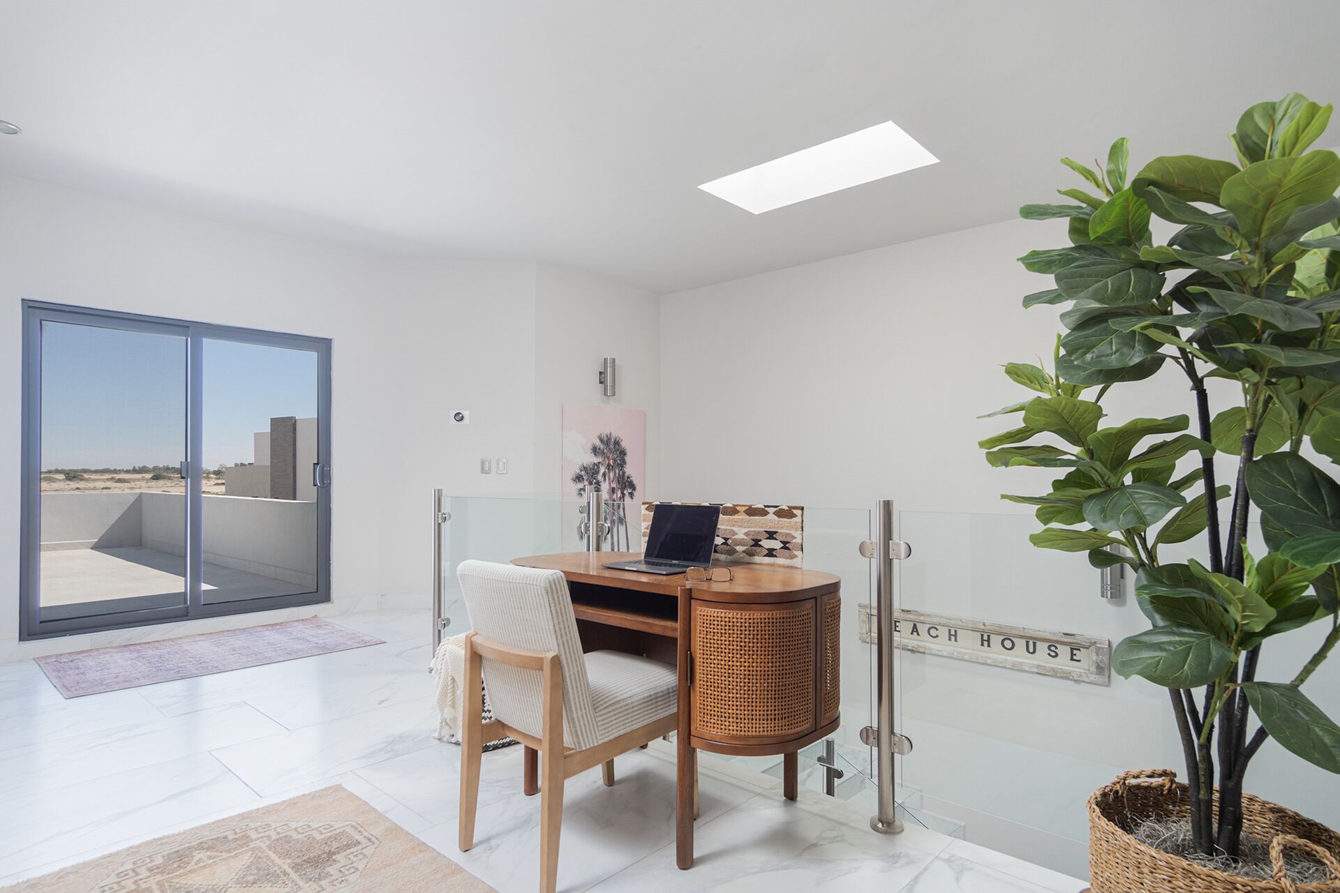 Un intérieur moderne avec une porte vitrée donnant sur un espace extérieur, un bureau en bois avec un ordinateur portable, une grande plante en pot et un panneau fixé au mur.