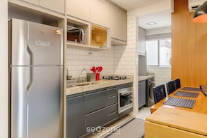 A modern kitchen with a stainless steel refrigerator, a microwave, and a built-in oven. There is a wooden dining table with chairs and a kitchen island with a sink and stove. - Cozy Apartment w Barbecue 550m - Beach Ire021 (Ubatuba)