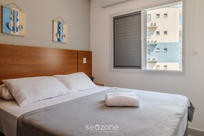 A neatly made bed with white linens and a gray blanket, a wooden headboard, and a window with blinds. - Cozy Apartment w Barbecue 550m - Beach Ire021 (Ubatuba)