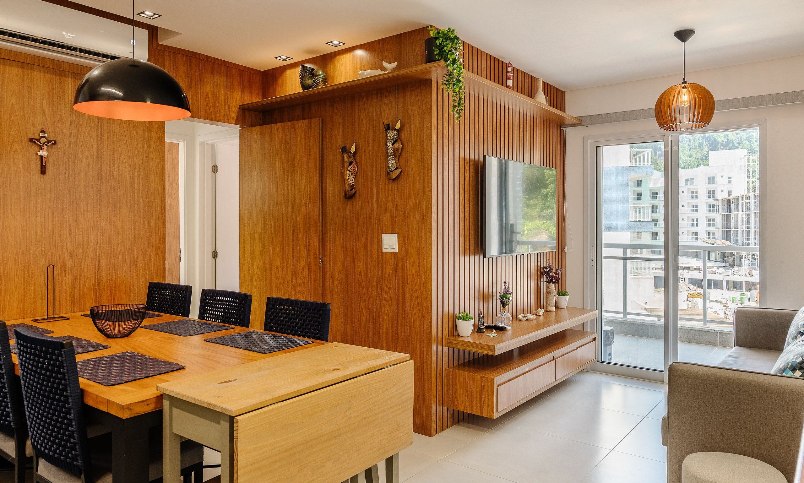Un comedor moderno con una mesa y sillas de madera, un televisor montado en un panel de madera, y un balcón con vistas a edificios.