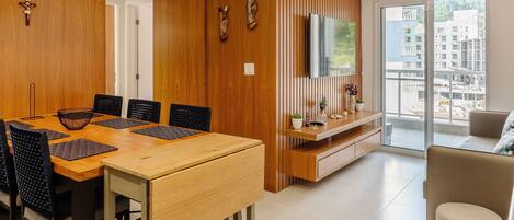 Un comedor moderno con una mesa y sillas de madera, un televisor montado en un panel de madera, y un balcón con vistas a edificios.