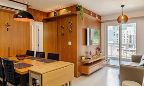 A modern dining area with a wooden table and chairs, a TV mounted on a wooden panel, and a balcony with a view of buildings. - Cozy Apartment w Barbecue 550m - Beach Ire021 (Ubatuba)