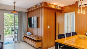 Un salón moderno con un televisor de pantalla plana montado en una pared de madera, una zona de comedor con una mesa y sillas de madera, y un balcón con vistas a zonas verdes.