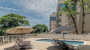 Un área de piscina en la azotea con sillones de descanso y sombrillas, rodeada por un rascacielos.