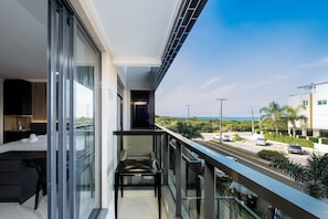 Un balcón moderno con puerta de cristal, una pequeña mesa y vistas a una calle con coches y palmeras.