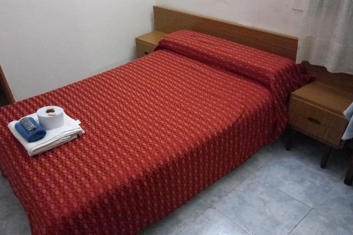 A single-bed room with a red patterned bedspread, a wooden nightstand, and a small drawer.