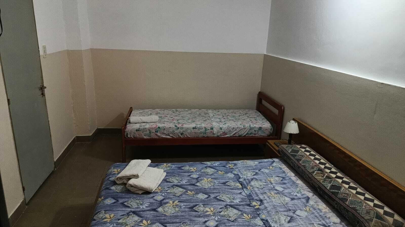 Een kleine, compacte kamer met twee eenpersoonsbedden, elk met een ander beddegoed. Er staat een klein tafeltje met een lamp naast een van de bedden.