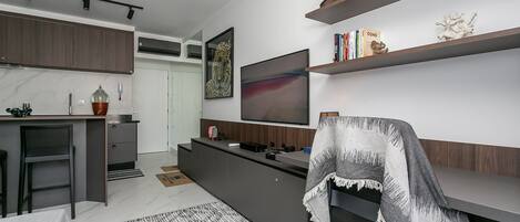 A modern kitchen with a built-in stove, a dining area with a table and chairs, a flat-screen TV mounted on the wall, and a bookshelf with books and decorative items.