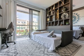 A modern bedroom with a large bed, a desk, and a shelving unit with decorative items. - Luxurious Studio Right on the Sand in Irb203 (Florianópolis)