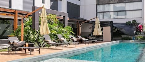 A poolside area with lounge chairs and umbrellas, a clear pool, and a modern building facade.