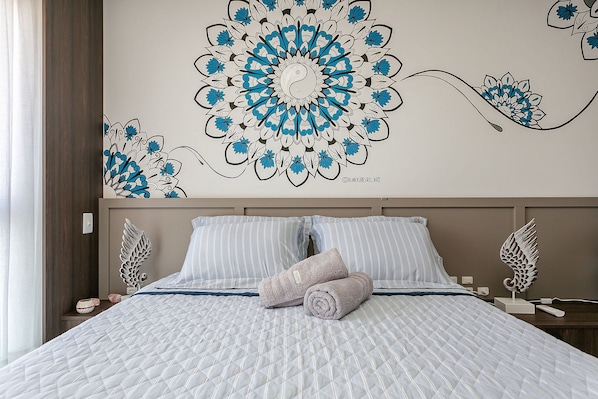 Un lit bien fait, recouvert d’une courtepointe blanche et bleue à motifs, avec une serviette enroulée et une œuvre d’art décorative accrochée au mur au-dessus.