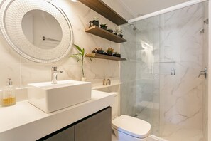 A modern bathroom with a white sink, a round mirror, and a glass-enclosed shower. - Luxurious Studio Right on the Sand in Irb203 (Florianópolis)