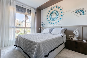 A neatly made bed with a quilted cover, a nightstand with a decorative item, and a wall with a large floral artwork.