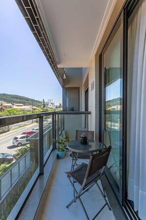 A balcony with a table and chairs, a potted plant, and a view of the street and hills. - Luxurious Studio Right on the Sand in Irb203 (Florianópolis)
