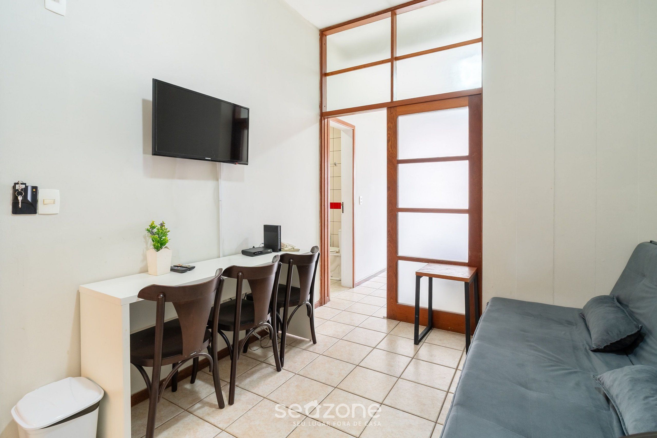 Un espacio habitable compacto con escritorio, sillas, televisor, sofá y puerta corredera de vidrio hacia un baño.