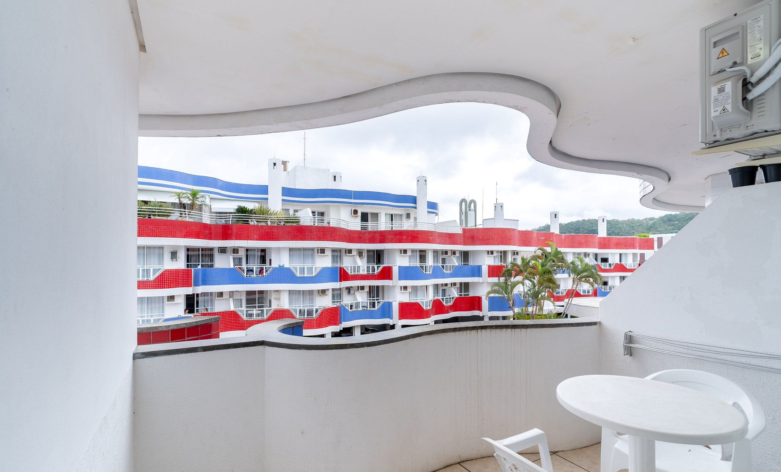 A balcony with a view of a multi-story building with balconies painted in red, white, and blue. There is a white table and two white chairs on the balcony.