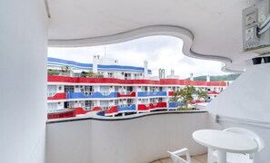 En balkon med udsigt til en bygning med flere etager, hvor balkonerne er malet i rød, hvid og blå. Der er et hvidt bord og to hvide stole på balkonen.
