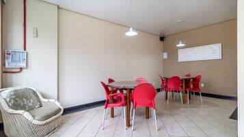 Un coin repas avec une table ronde et des chaises rouges, un canapé en osier et un panneau électrique fixé au mur.
