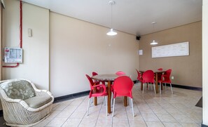 Un coin repas avec une table ronde et des chaises rouges, un canapé en osier et un panneau électrique fixé au mur.