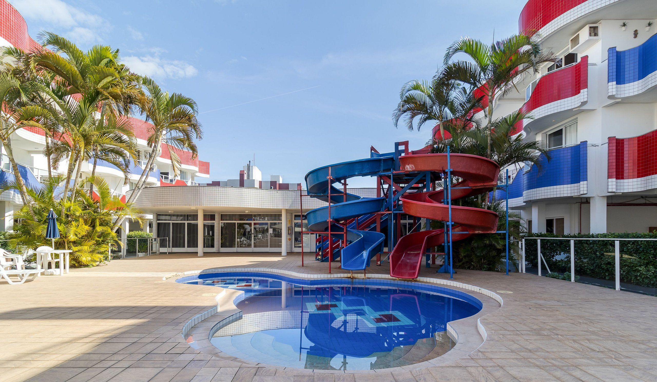 Zona de piscina con tobogán de agua, palmeras y un edificio con balcones rojos y blancos.