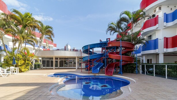 Ein Poolbereich mit Wasserrutsche, Palmen und einem Gebäude mit roten und weißen Balkonen.