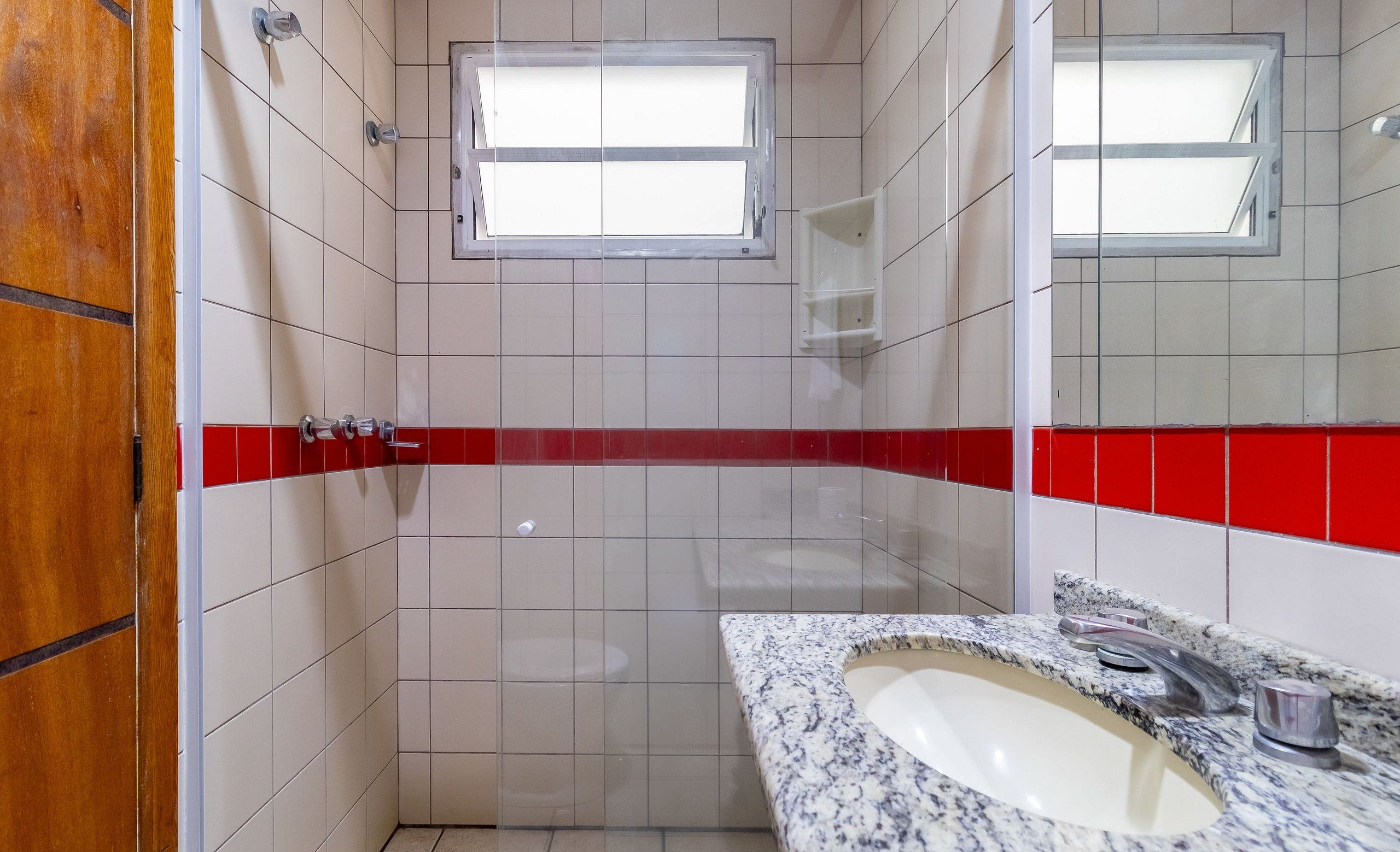 A bathroom with white tiled walls, a red accent strip, a window, a sink with a granite countertop, and a mirror.