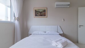 Una cama bien hecha con sábanas blancas y una toalla enrollada, un cuadro en la pared y un equipo de aire acondicionado.