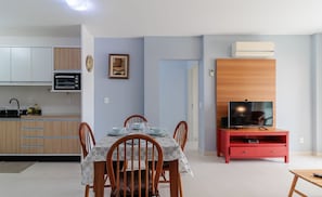 A modern kitchen with white cabinets, a built-in oven, and a microwave. There is a dining area with a table set for four, surrounded by wooden chairs. A television is placed on a red stand next to a wooden cabinet.