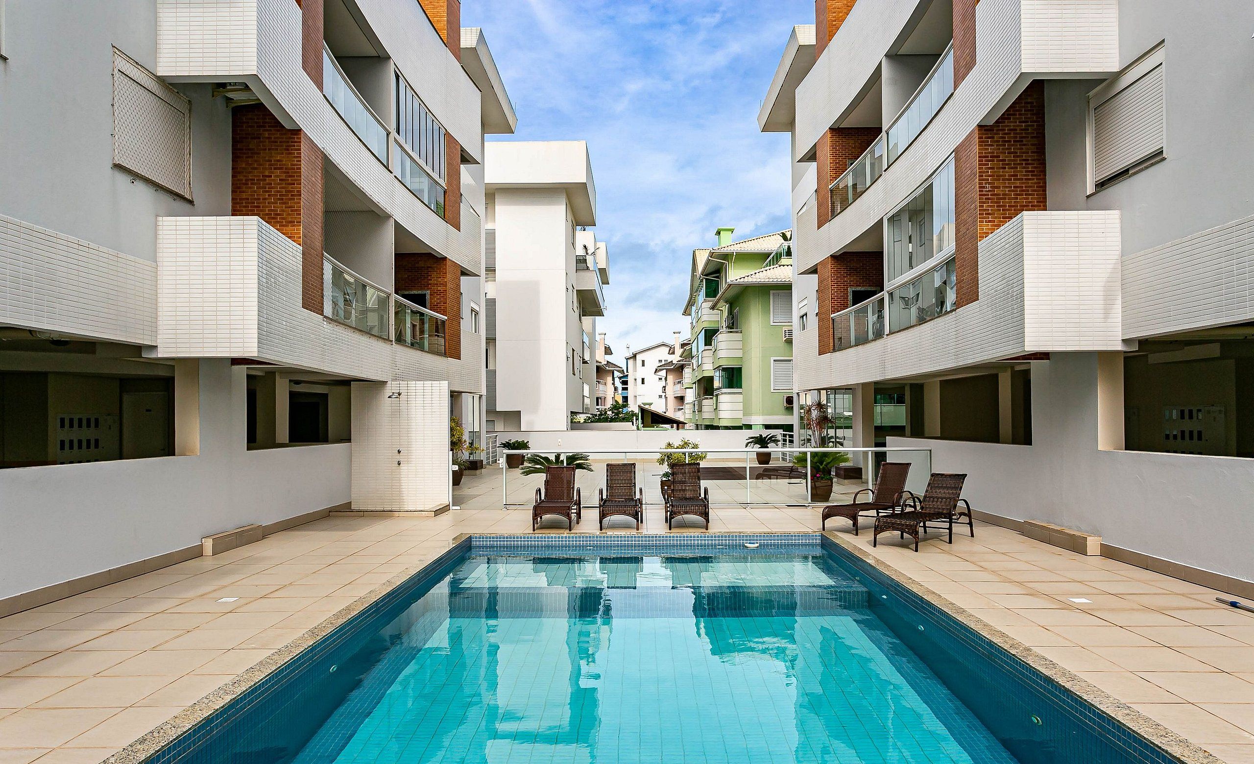 Una piscina rodeada de edificios modernos con balcones, cielo despejado y algunas sillas de descanso.