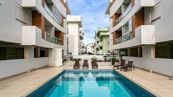 Una piscina rodeada de edificios modernos con balcones, cielo despejado y algunas sillas de descanso.