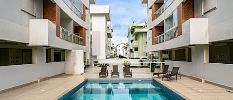 A swimming pool surrounded by modern buildings with balconies, a clear sky, and a few lounge chairs.