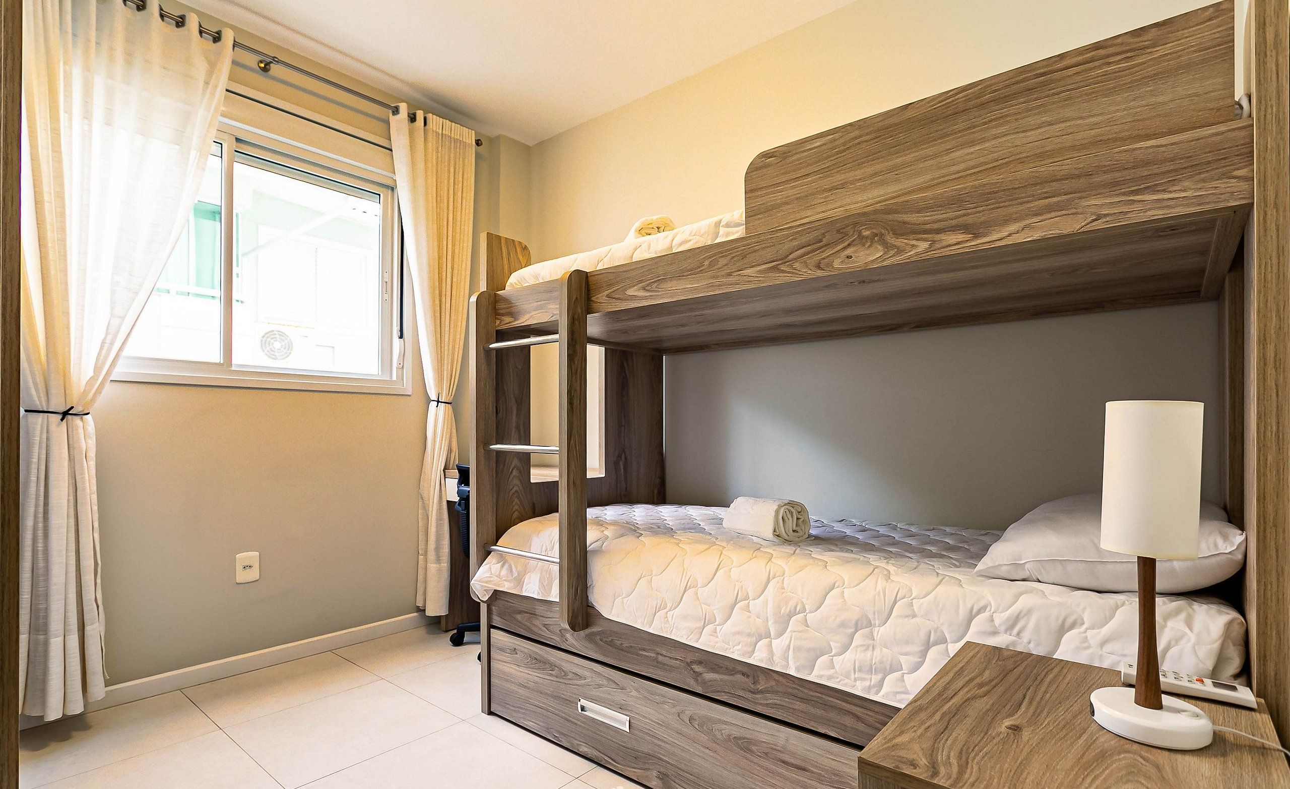 Una cama litera con cabecera de madera, una mesita de noche con lámpara y una ventana con cortinas.