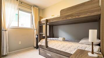 Uma cama de solteiro com cabeceira de madeira, uma mesinha de cabeceira com abajur e uma janela com cortinas.