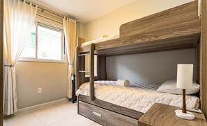Uma cama de solteiro com cabeceira de madeira, uma mesinha de cabeceira com abajur e uma janela com cortinas.