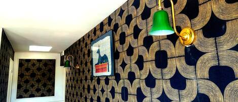 A hallway with patterned wallpaper, a framed picture, and wall-mounted lamps.