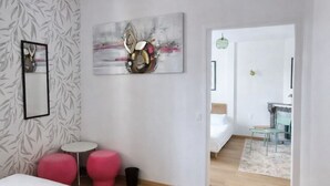 Habitación de hotel moderna con una cama grande, una pequeña mesa redonda, dos taburetes rosa, una obra de arte abstracta en la pared y un baño visible al fondo.