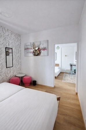 Habitación de hotel moderna con una cama grande, una pequeña mesa redonda, dos taburetes rosa, una obra de arte abstracta en la pared y un baño visible al fondo.